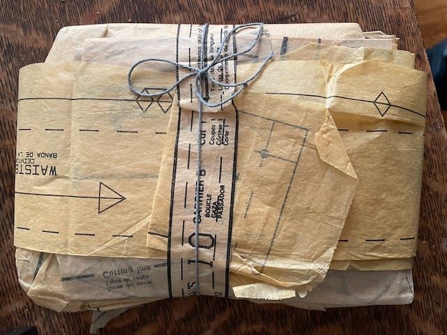 An unbranded parcel wrapped in printed sewing pattern tissue parts + tied with a cotton cord, sitting on a wooden surface
