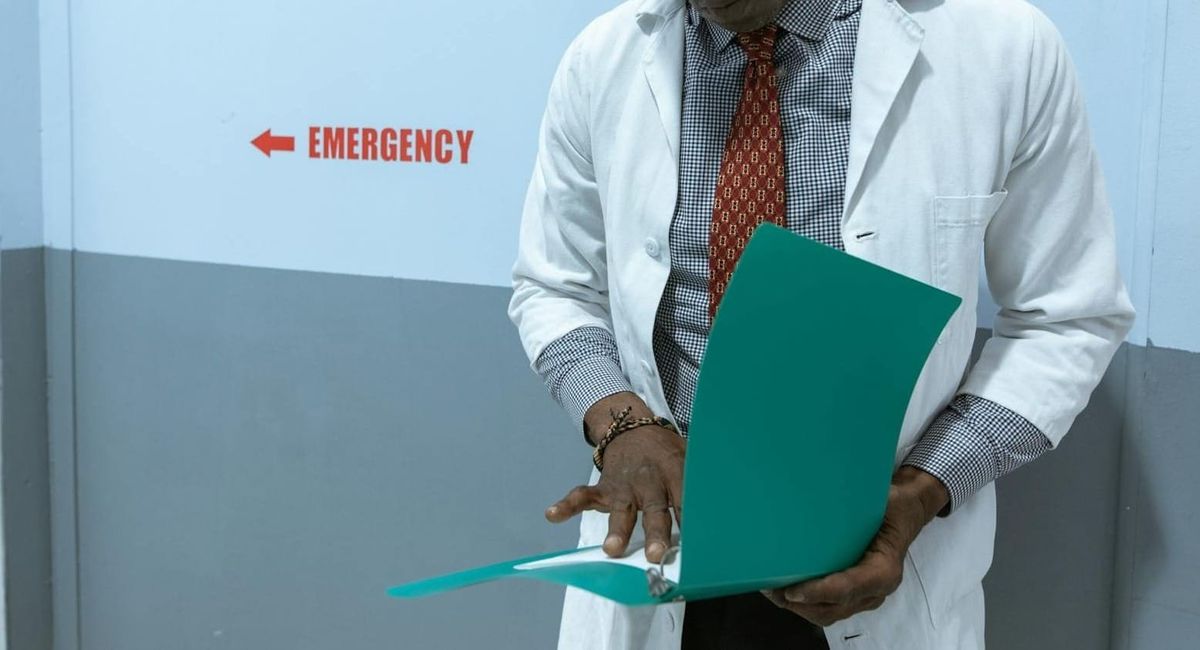A doctor in a lab coat examines documents near the emergency room sign.