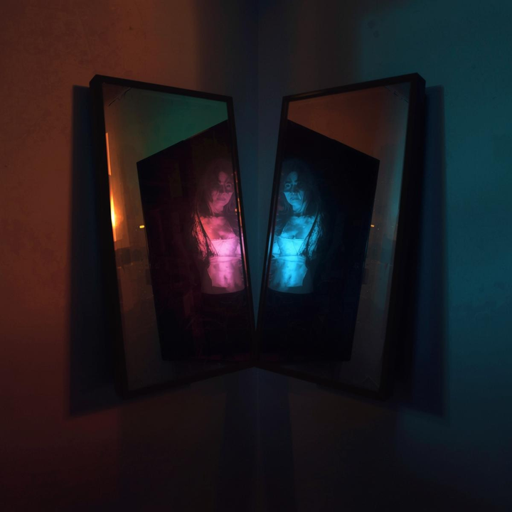 Two mirrors in a dim corner reflecting the same figure in warm red and cool blue light.
