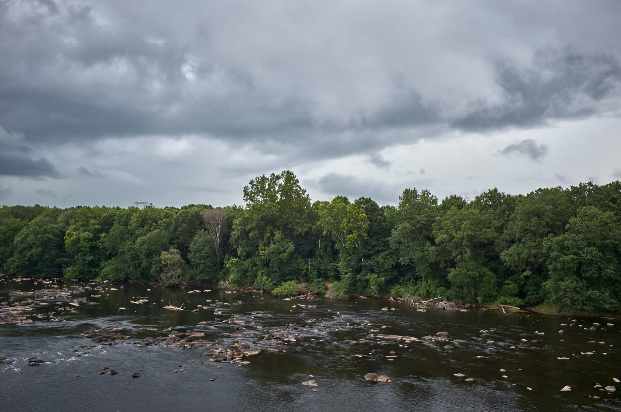 The view of a rock filled river and the forested banks with dark clouds filing the sky.