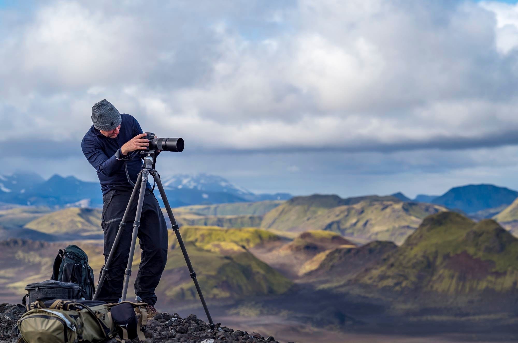 David looking through a camera on a tripod at the top of a mountain in Iceland with green and blue mountains in the background and a white cloudy sky.