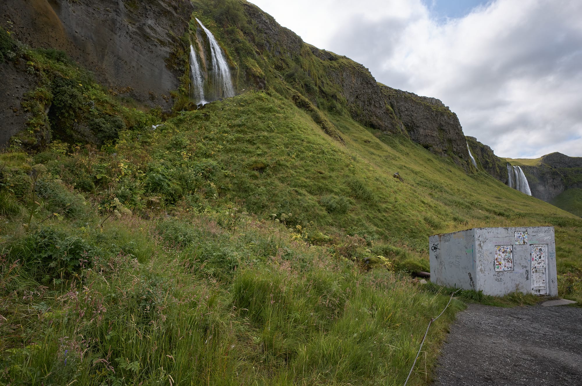 Two waterfalls on a green hillside with a grey building full of graffiti.