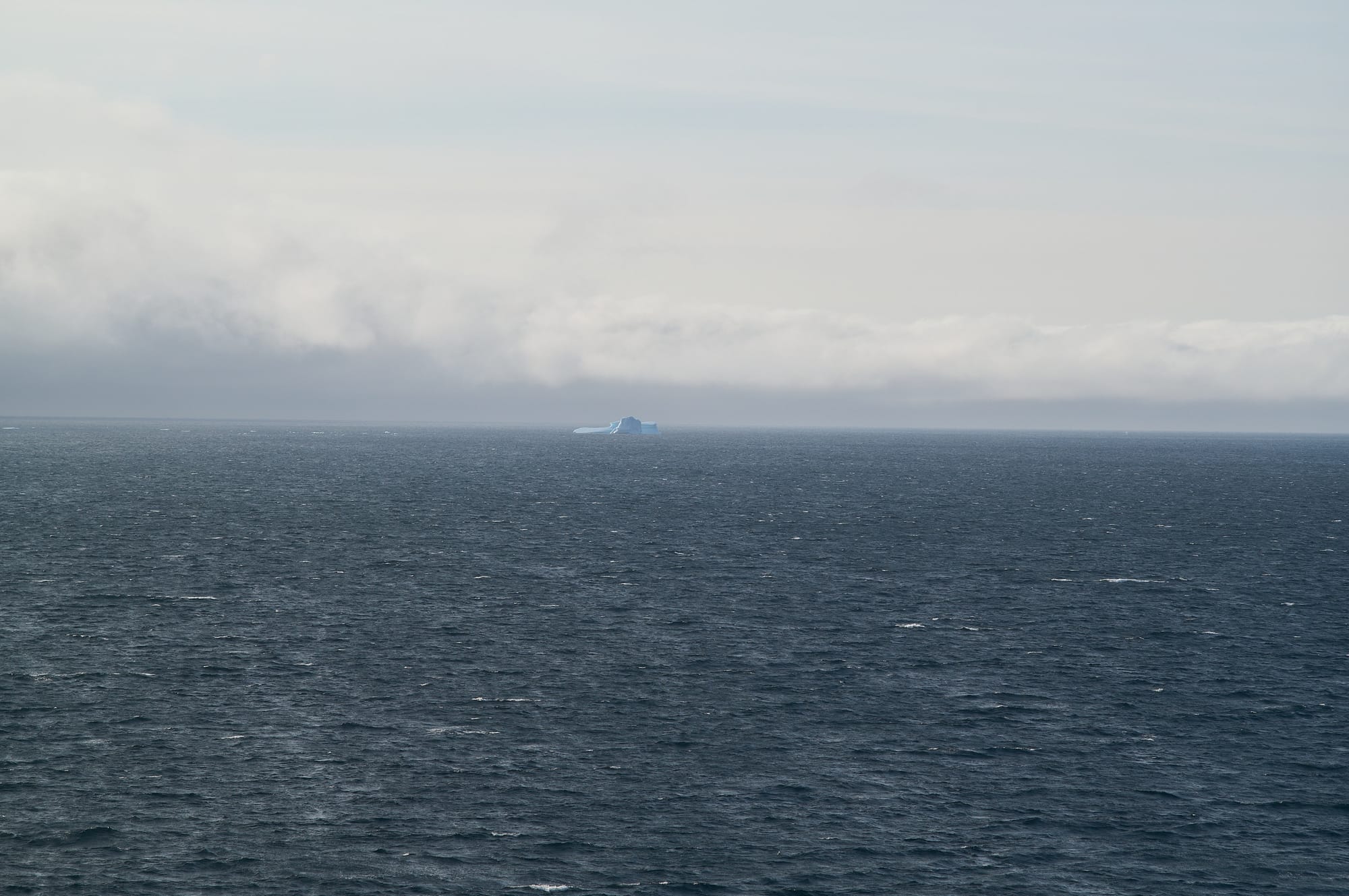 Ocean view with clouds and a solitary iceberg in the distance.