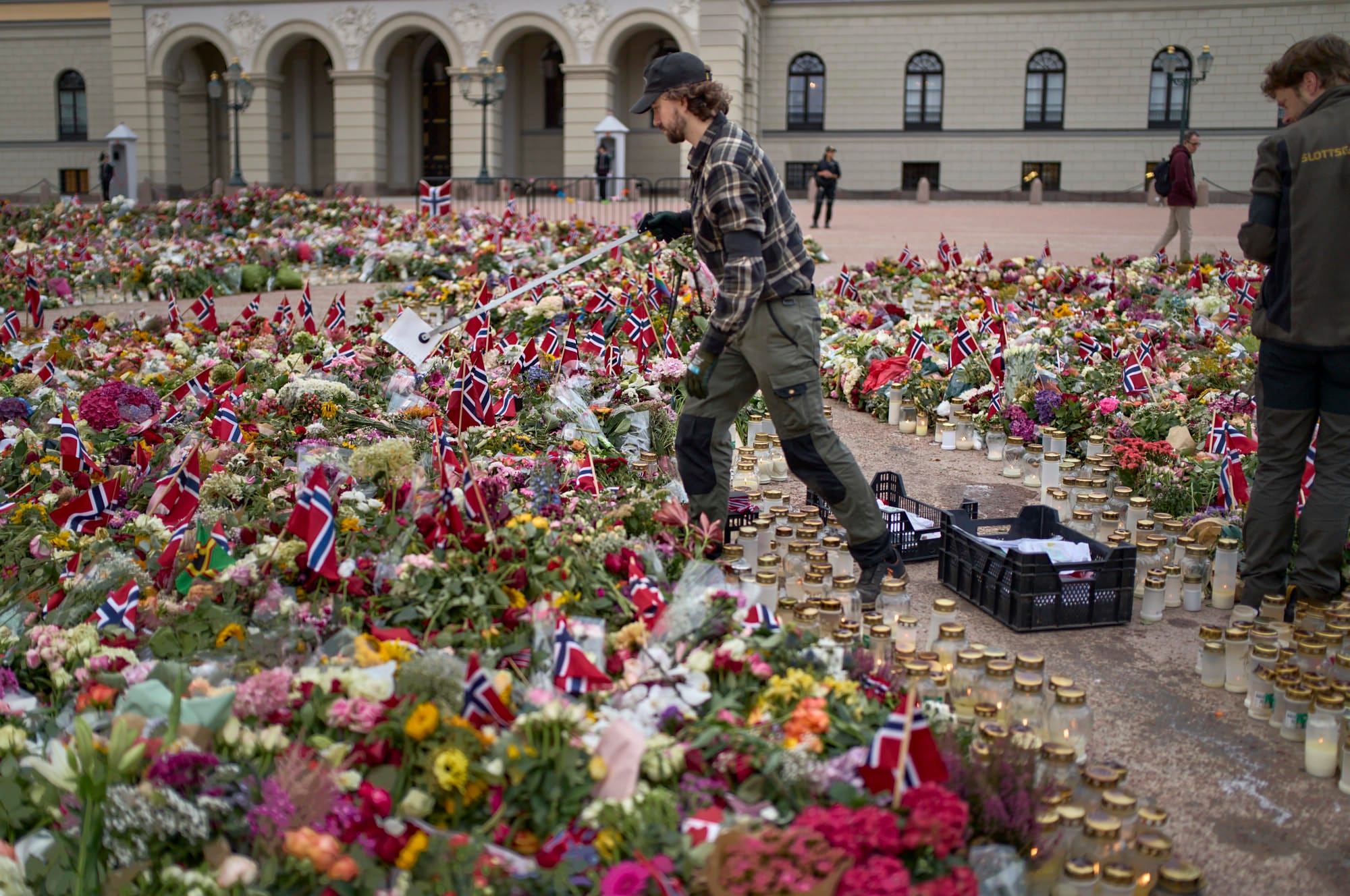 Man with long tongs picking up paper in the middle of flowers, flags and candles.