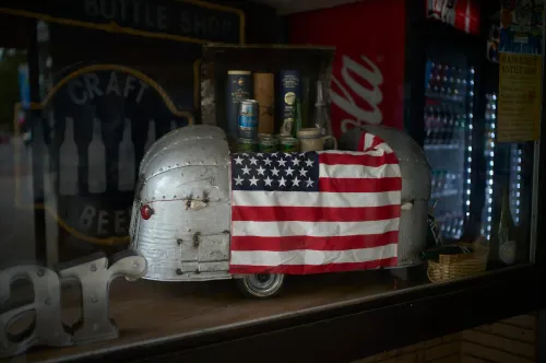 A retail window with a model airstream and a large american flag draped over the top with bottles in the background.
