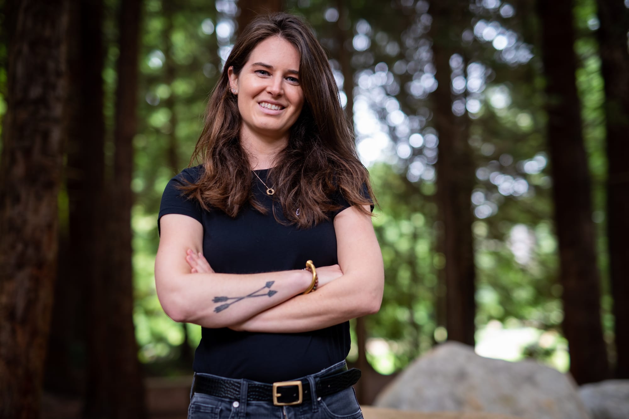 Person with long brown hair smiles down at the camera with arms crossed showing a tattoo with two crossed arrows. In the background are redwood trees, rocks, and greenery.