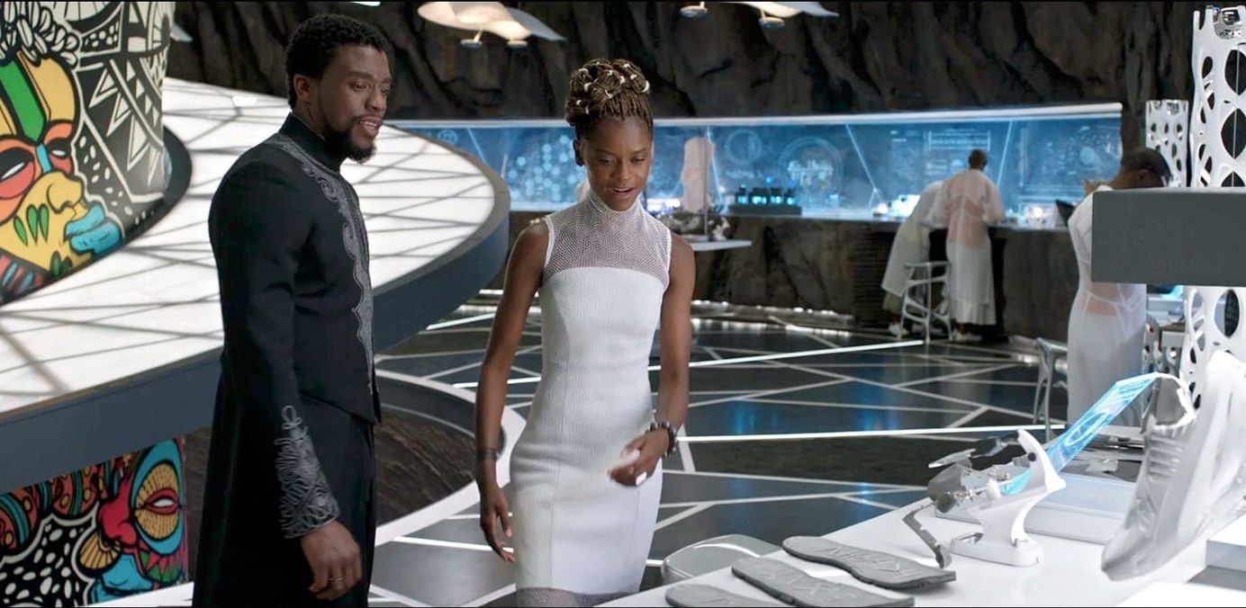 Black Panther and his sister, Shuri, stand In a lab with tech equipment and african murals on the wall. 