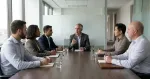 Six people are sitting around a boardroom table, only one person at the table talking. 