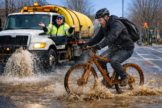 Cyclist splashed by brine while riding through pooled liquid from repeated bike-lane treatment on a dry winter day.