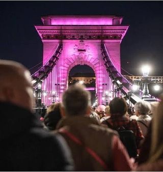 People gather before the pink-lit stone tower of Budapest’s Chain Bridge at night.