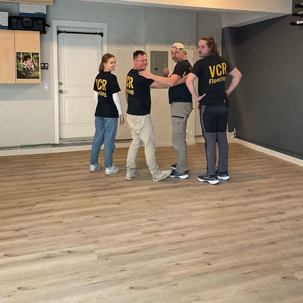 A group of four people wearing matching "VCR Flooring" shirts stand in a finished garage with new wood-look flooring, smiling and posing for the photo.