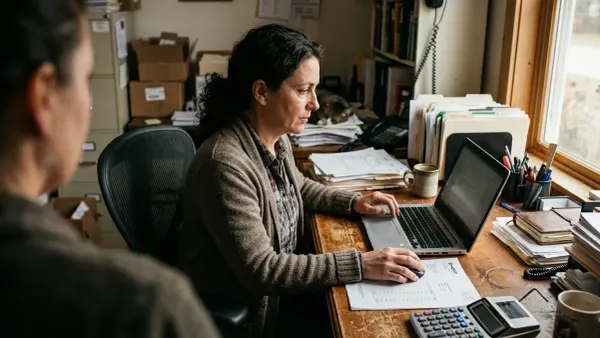 A small business owner sitting at a desk with a laptop, an invoice, and an adding calculator in front of her.