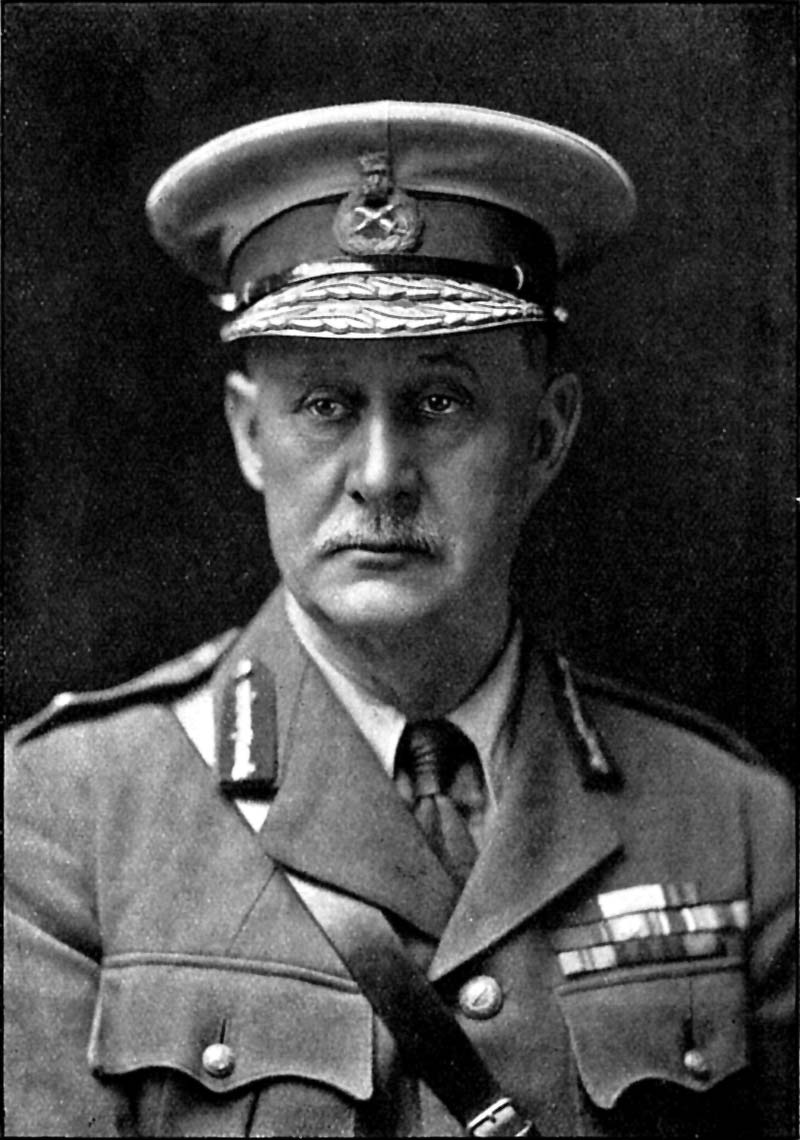 A black and white formal portrait photograph shows a middle-aged man wearing a military uniform. He has a neatly groomed mustache and is looking directly at the camera with a serious expression. The subject wears a peaked military cap with decorative braiding around the visor and what appears to be a badge or emblem on the front. His uniform jacket features shoulder boards with rank insignia, collar tabs, and multiple ribbon bars or medal ribbons arranged in rows on the left chest area. The uniform appears to be of a formal military style with buttons visible down the front and various decorative elements typical of high-ranking military dress. The photograph is taken against a dark, plain background, creating a formal portrait composition typical of official military photography. The image quality and style suggests it was taken in the early-to-mid 20th century. (ALT Text by Claude AI)
