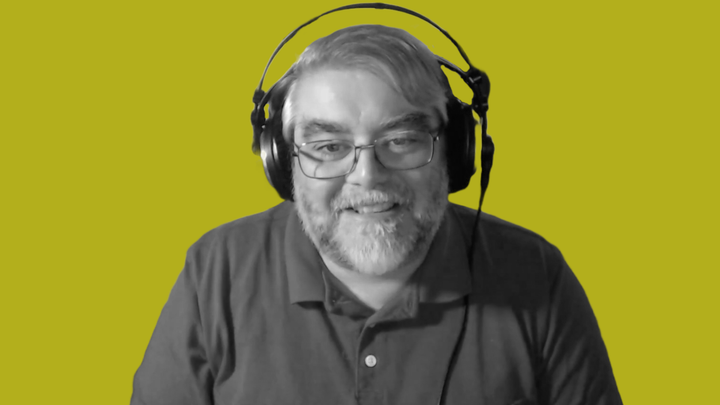 Raph Koster with a headset on, smiling at the camera. He's black & white in front of the grokludo yellow background.