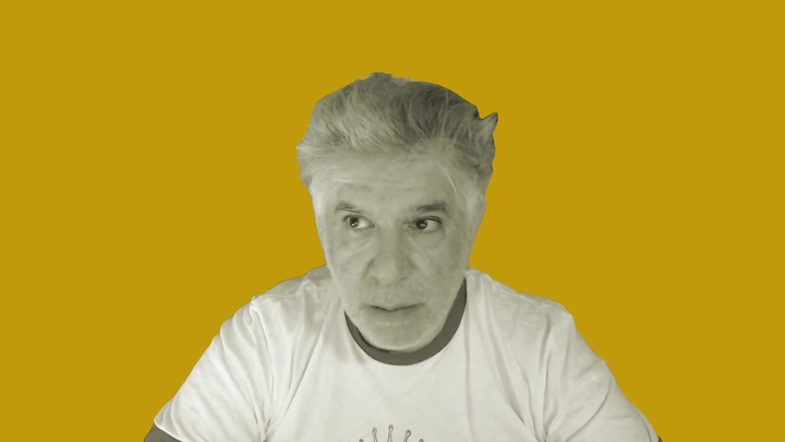 Nikita Mikros reflects on Killer Queen as a guest on grokludo. He is seen in black and white, from the chest up, in front of an orange background.