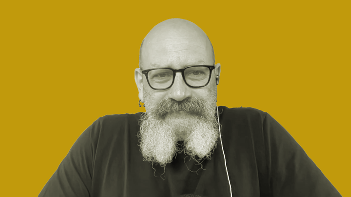 Seán Dagher in glasses and a black shirt in front of an orange background, talking abotu sea shanties on the grokludo podcast.