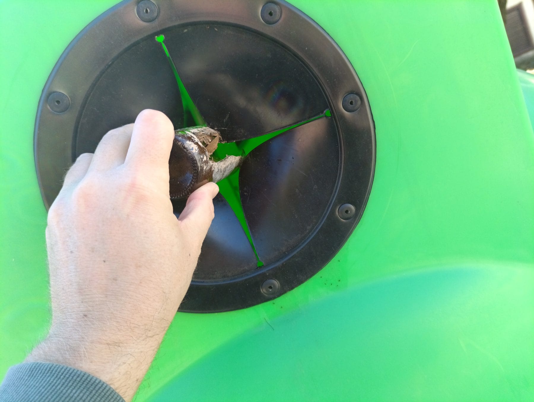 Mano introduciendo una botella de vidrio rota por la abertura circular con solapas negras de un contenedor de vidrio verde, vista cercana
