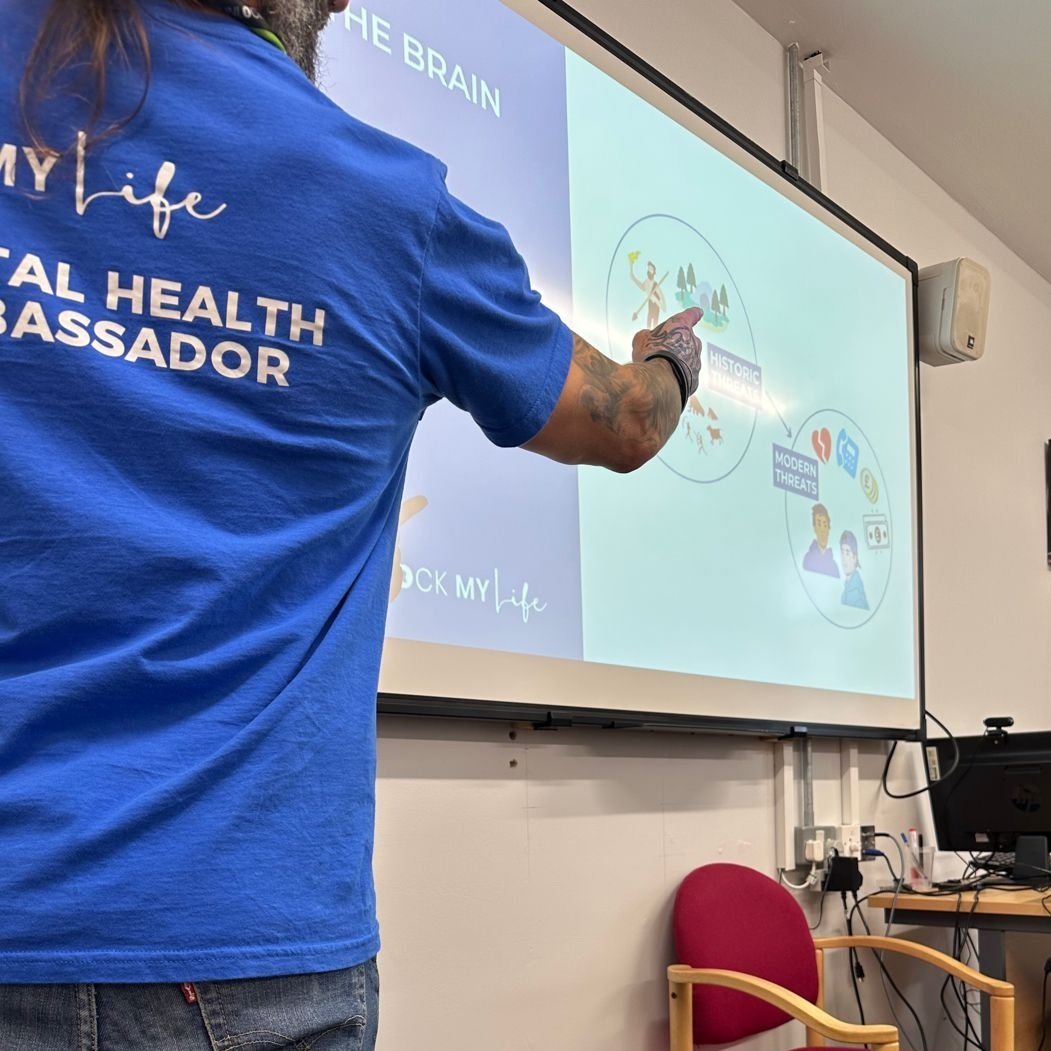 A man wearing a 'Mental Health Ambassador' t-shirt points to a slide during a BrainSmart training session, showing diagrams about threat responses.