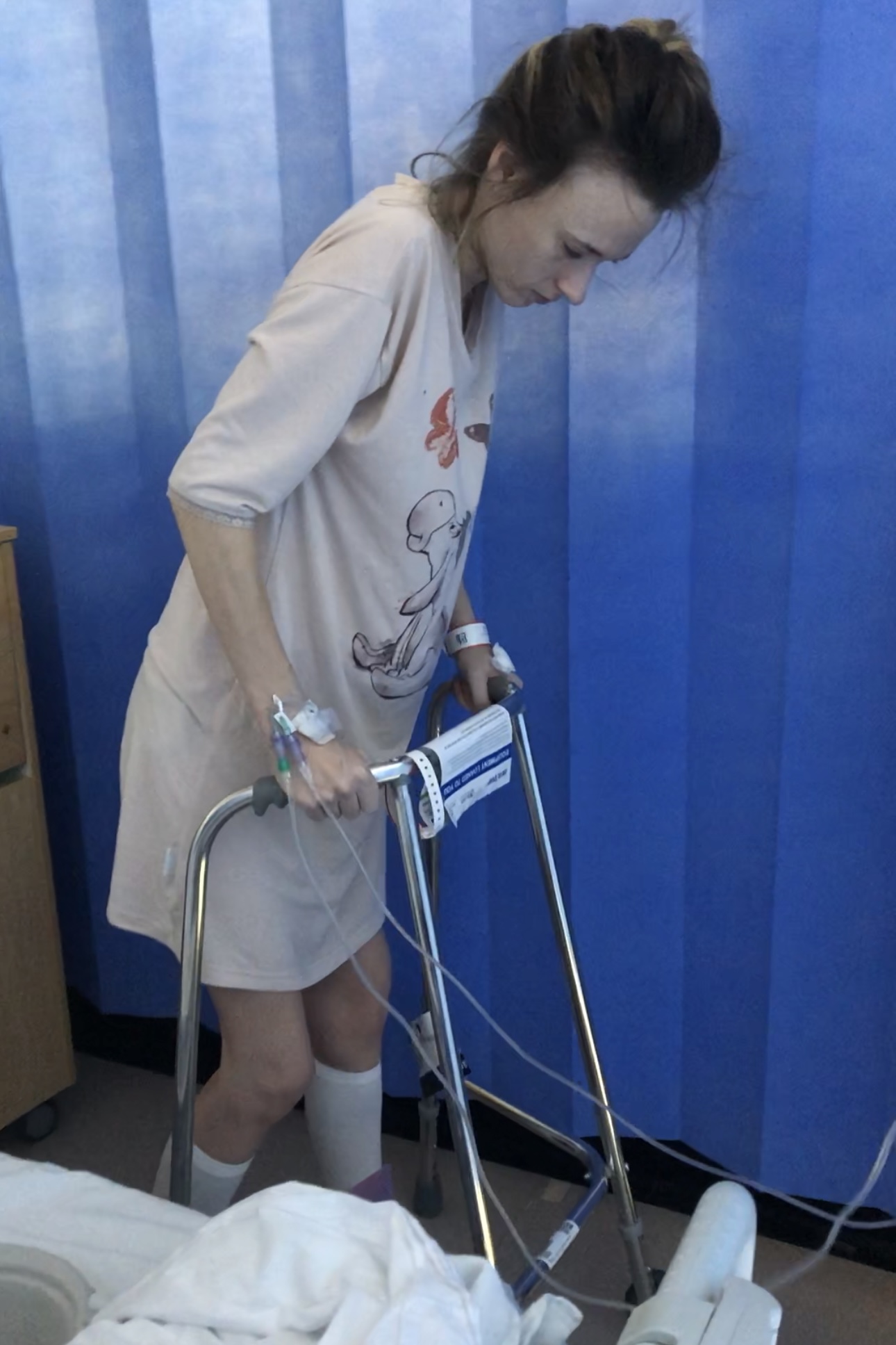 Monica Thomas using a walking frame in a hospital room, wearing a medical gown with IV tubes in her arms, standing beside a blue privacy curtain
