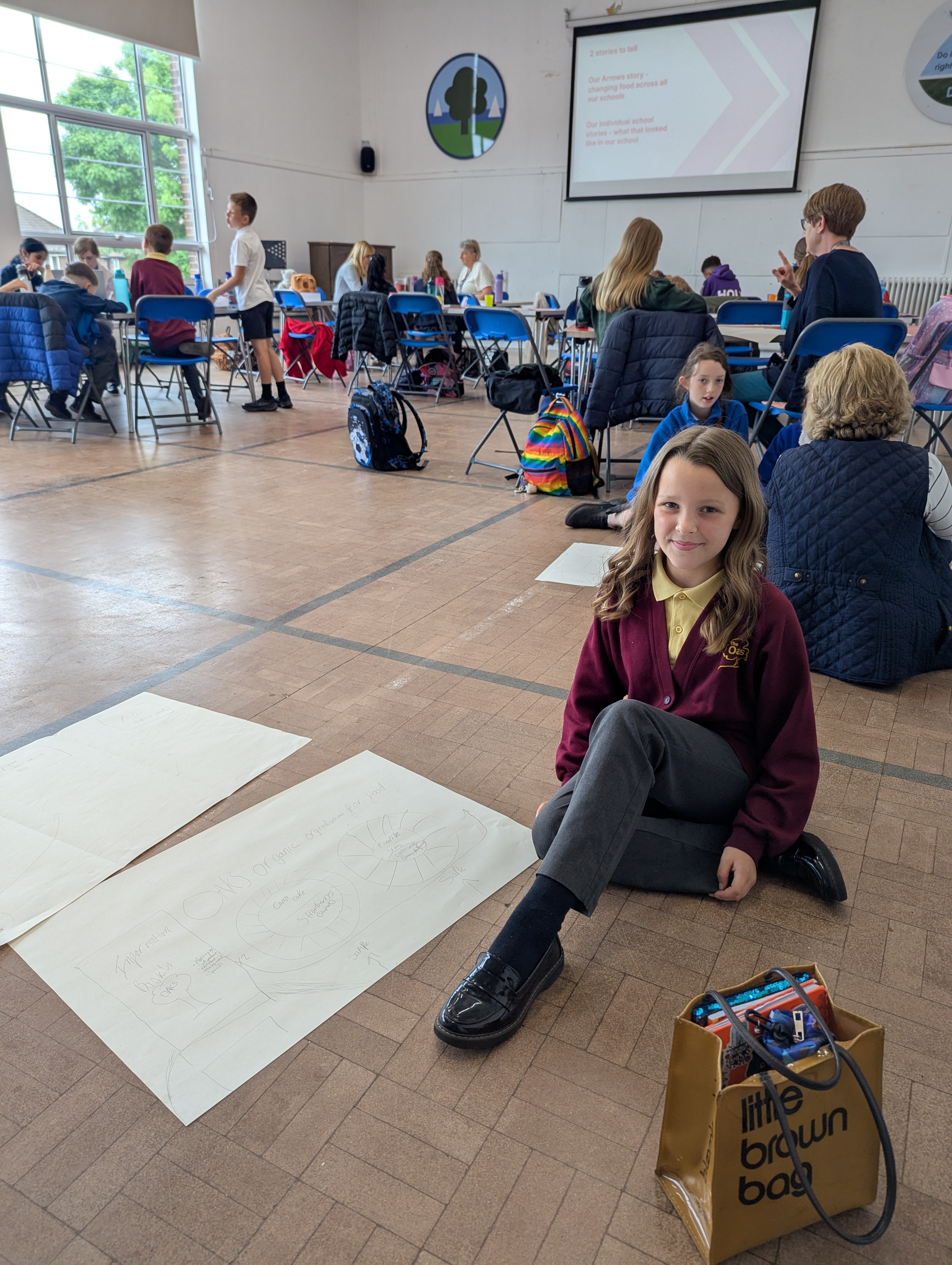 A primary school pupil sits next to a hand-drawn poster on composting and snacks at a school hall event, smiling with a gold ‘Little Brown Bag’ beside her