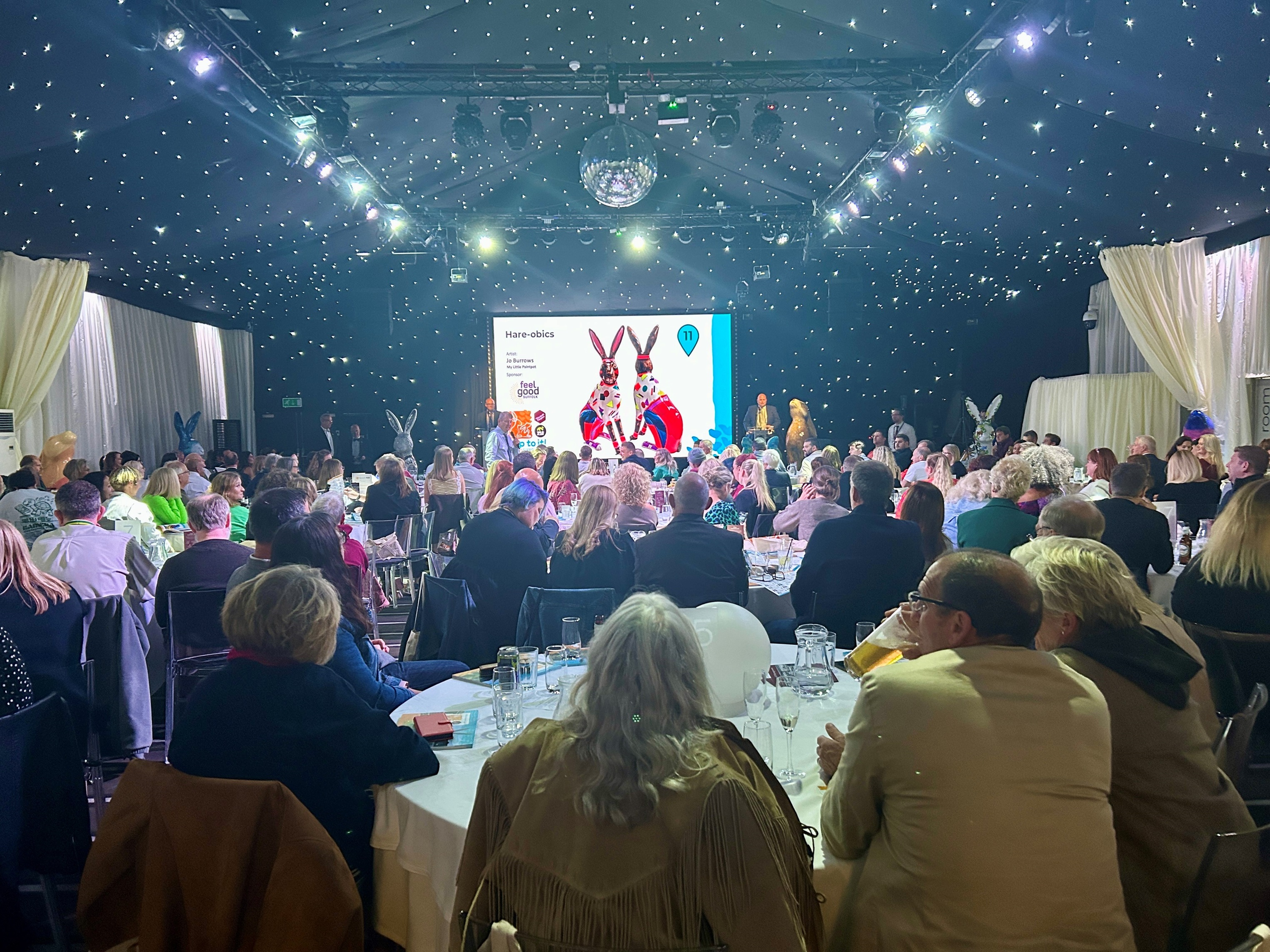 Large crowd seated at tables watching the stage during the hospice hare auction, with sculptures visible in the background
