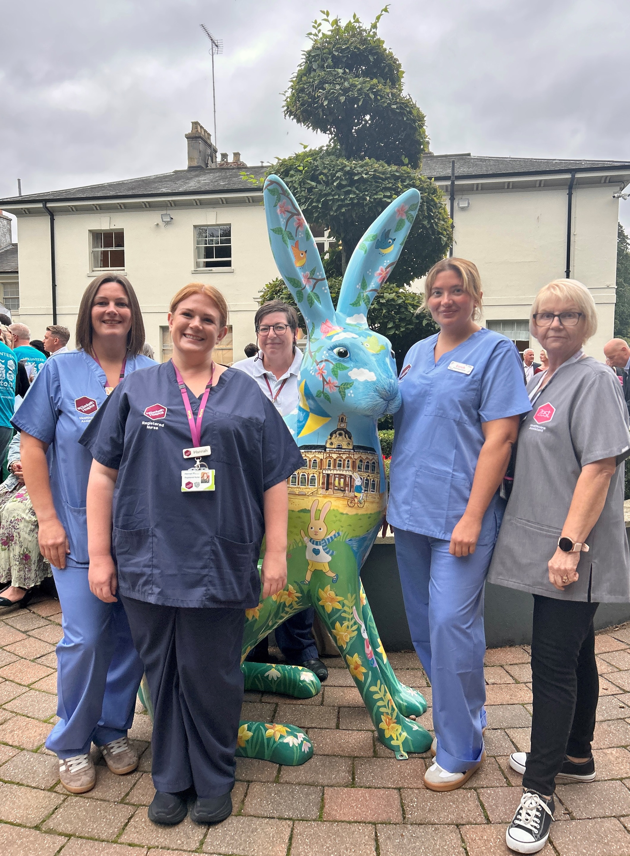 Five hospice nurses standing outdoors beside a large blue hare sculpture decorated with Ipswich landmarks and clouds