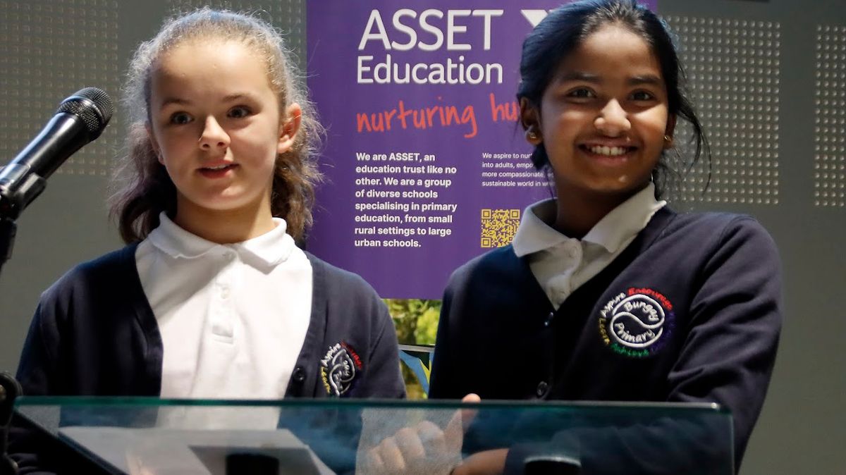 Two primary school pupils in blue uniforms speak from the stage in front of an Asset Education banner