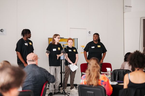 Three young people in black "The Power of Youth" t-shirts stand at a microphone presenting to a seated audience in a white-walled room
