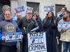 Attorney Adam White speaks into a microphone at a podium during a press conference surrounded by people holding signs of obsc