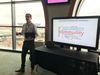 A facilitator stands in front of a flat screen TV with a word cloud, and speaks to participants at a "Visioning session" at C