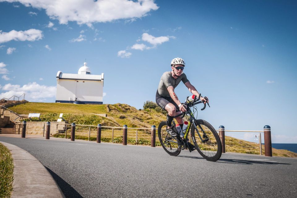 Campbell Wallace riding a road bike around a coastal bend past a small white lighthouse