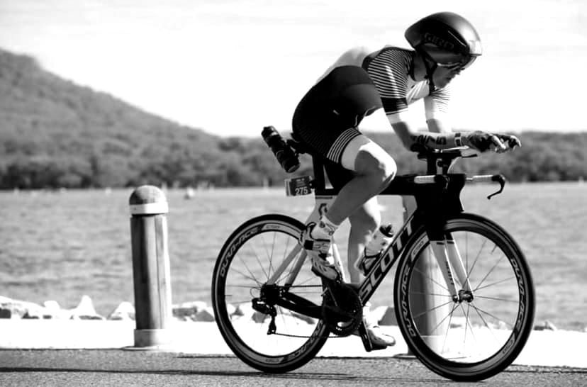 Black-and-white shot of David Bones riding a time-trial bike along a waterfront road
