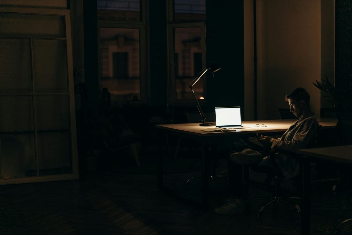 Person sitting at desk in dark room with laptop and desk lamp, representing willpower depletion and mental fatigue