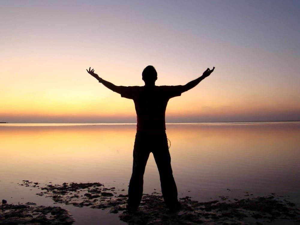 A person standing at the water's edge with arms wide open against a warm golden sunset — symbolizing resilience, freedom and rebuilding after adversity