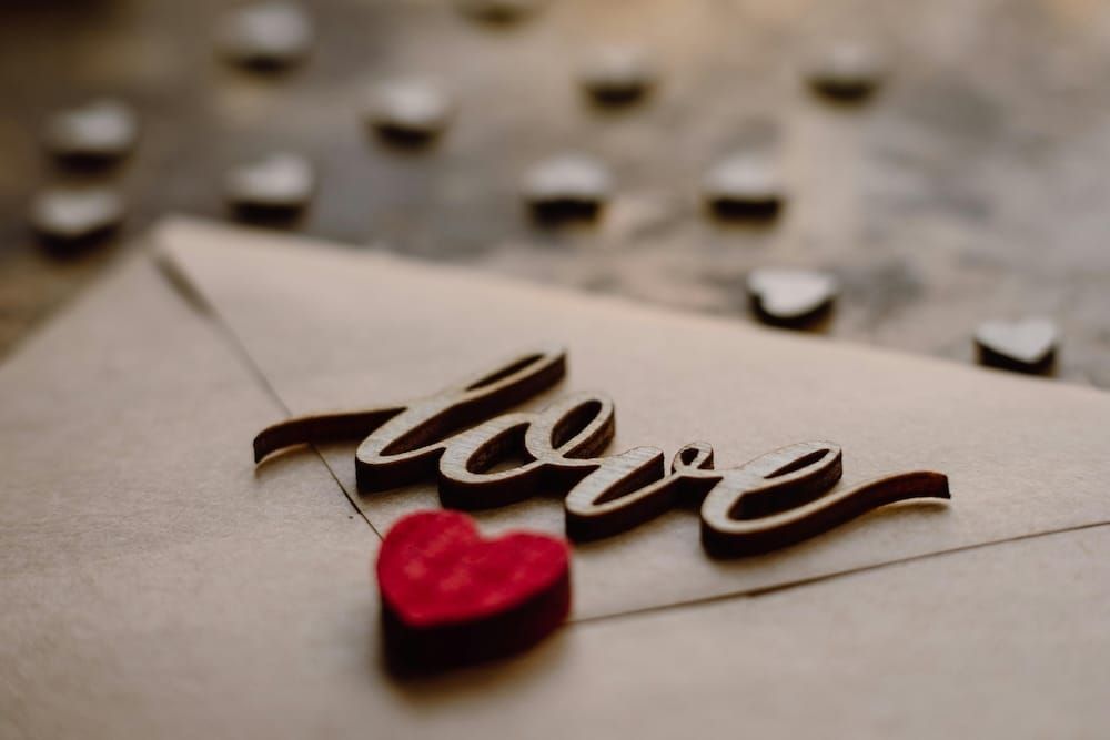 木質雕刻的「love」字樣放在信封上，旁邊散落著小木心，前景有一顆紅色絨毛心形裝飾，背景淺焦模糊，整體色調溫暖復古。