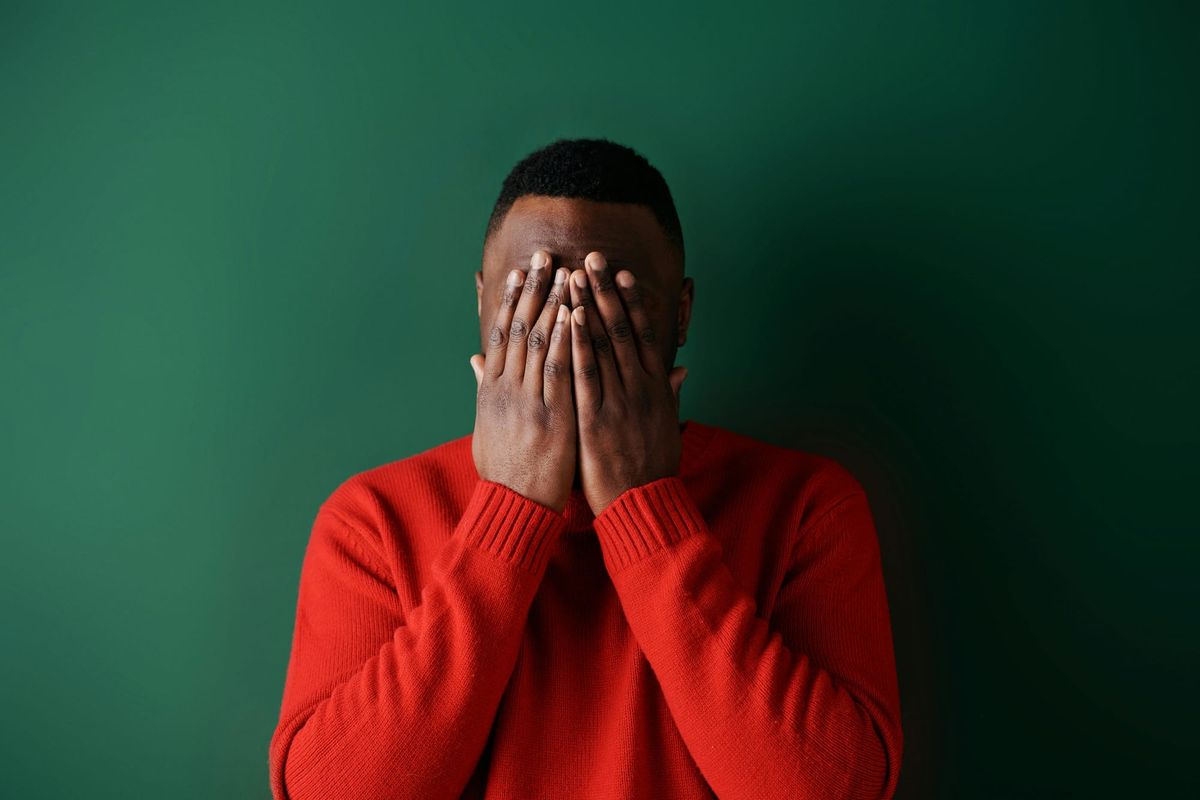 A man in a red sweater covering his face with both hands against a dark green background, expressing shame or distress.