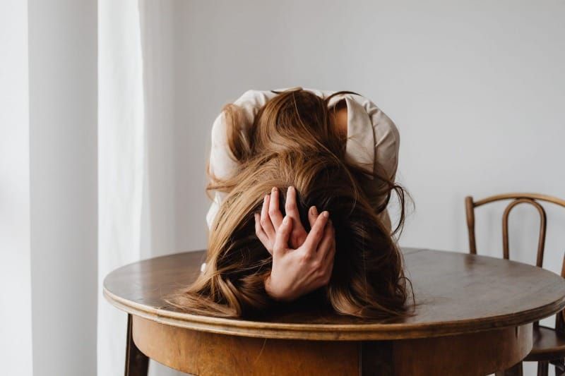 A woman rests her head face-down on a wooden table, hands in her hair, looking exhausted.