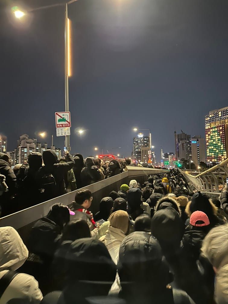 어두운 밤, 한강의 큰 다리의 인도에 겨울옷을 입은 사람들이 꽉 차서 한 방향으로 이동하고 있다.