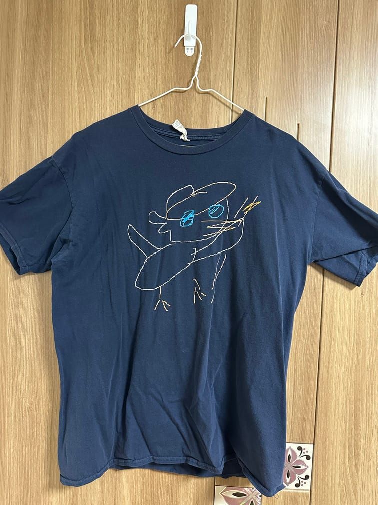남색 티셔츠의 앞면에 그림판으로 그린 것처럼 도트가 노출된 선으로 모자를 쓰고 막대기를 들고 있는 오리가 그려져 있다.