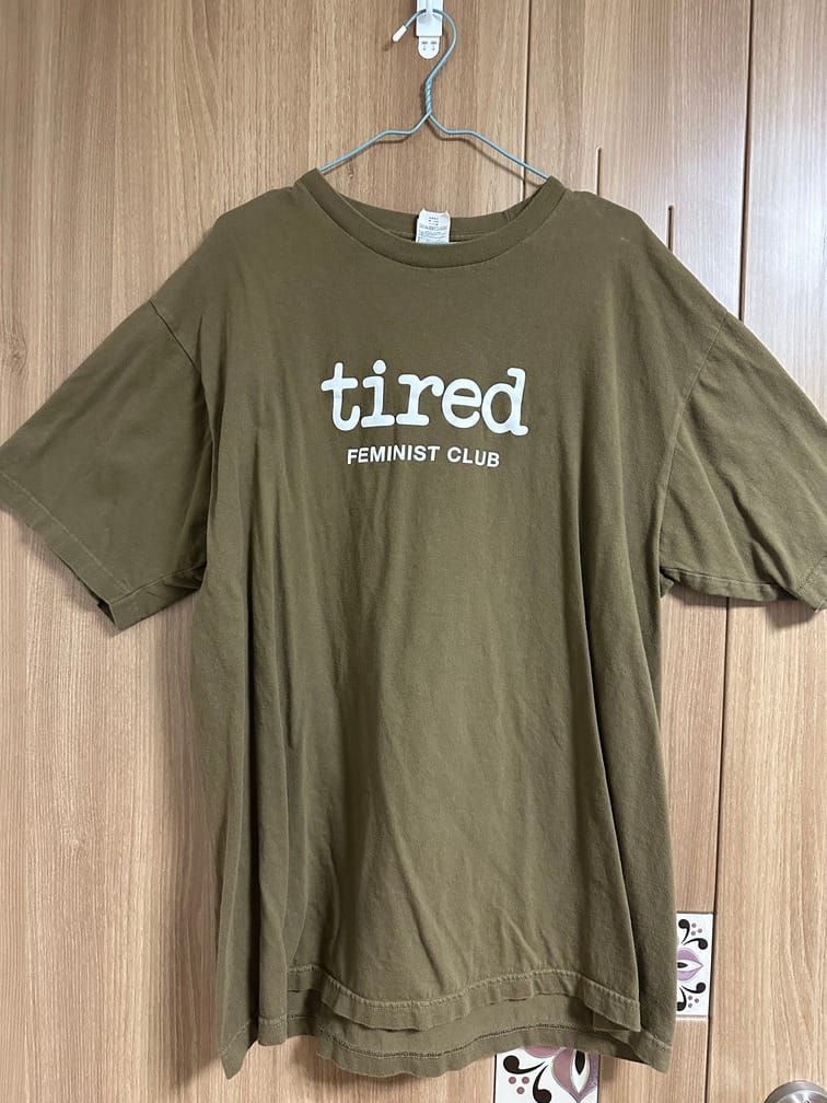 카키색 반팔 티셔츠의 가슴에 "tired FEMINIST CLUB"이라고 흰 글씨로 적혀 있다.