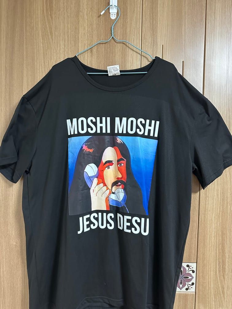 검은 티셔츠의 가운데에 MOSHI MOSHI JESUS DESU라는 흰 글자와 함께, 장발과 수염의 선해보이는 남성이 수화기를 든 모습이 애니메이션풍으로 묘사되어 있다.