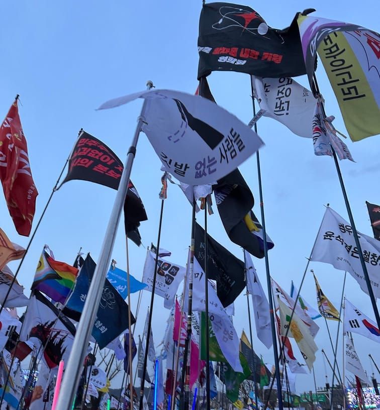 오후 하늘을 배경으로 수십여개의 깃발이 보이는 사진을, '깃발 없는 사람들'이라는 깃발을 들고 있는 사람의 시점에서 찍은 사진.