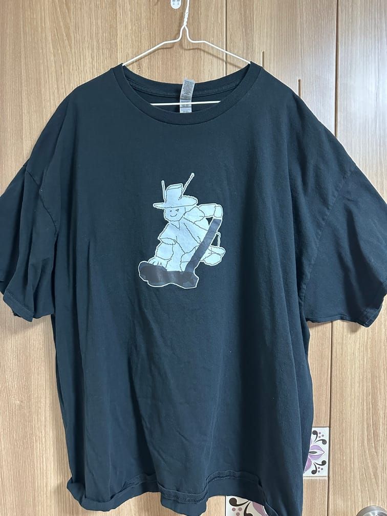 검은 티셔츠의 가운데에 흰 배경에 선으로 묘사되어 갓을 쓰고, 검은 무언가를 펴바르고 있는 듯한 사람의 도안이 그려져 있다.