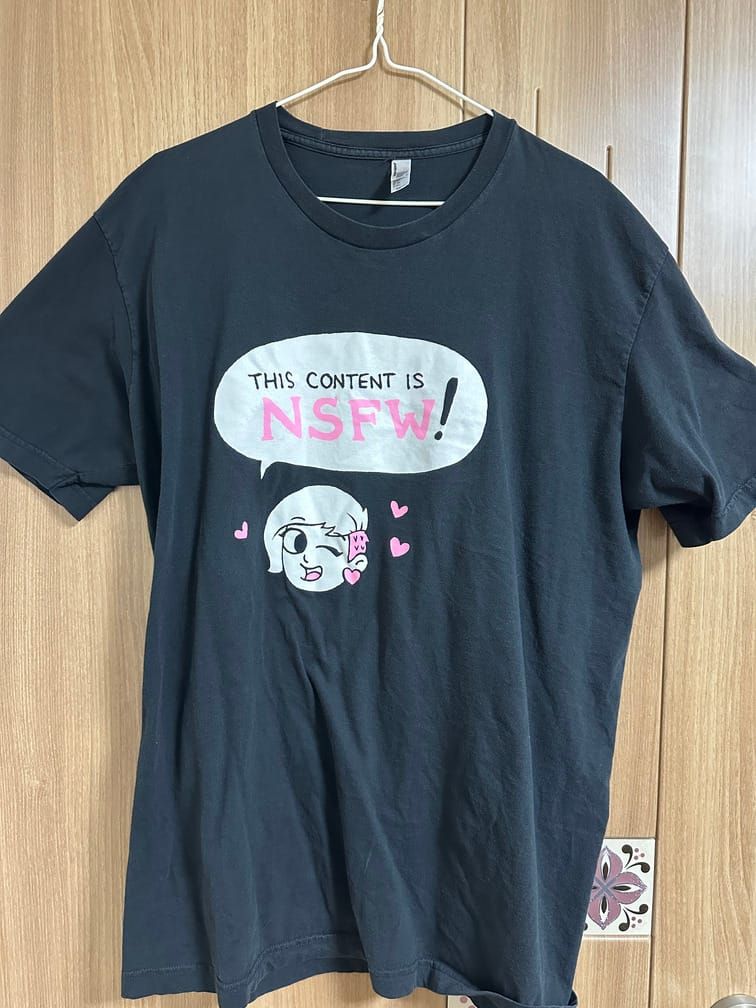 검은 티셔츠의 가운데에 투블럭 컷을 한 여성 캐릭터가 윙크하며 말풍선으로 "This content is NSFW!"라고 말하는 모습이 중간중간 핑크색 포인트와 함께 들어간 모습.