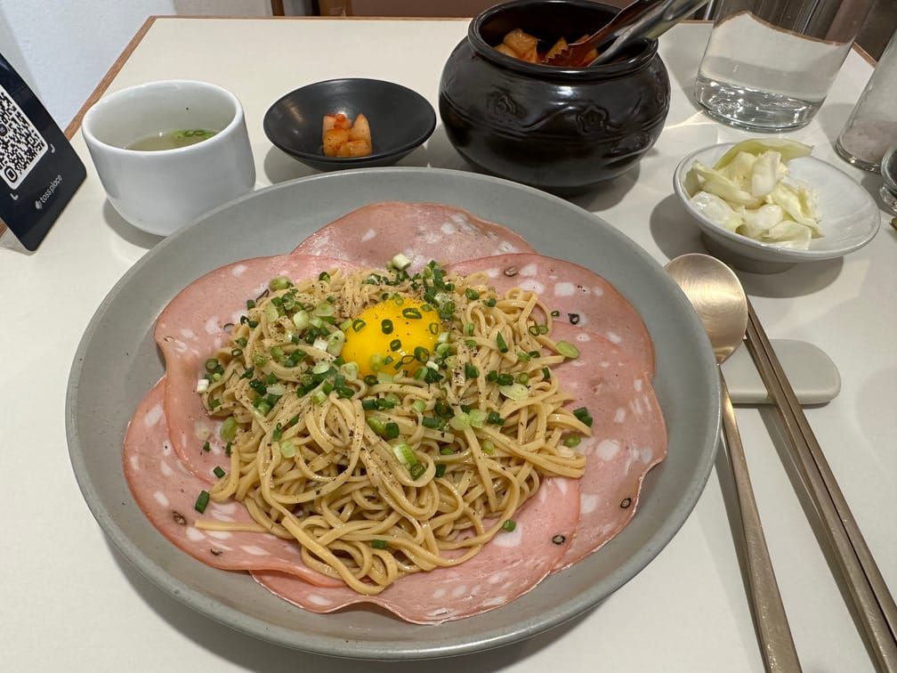 그릇에 햄이 깔려 있고, 그 위에 면이 차려져 있고 쪽파와 후추, 달걀 노른자가 올라가 있다. 김치, 양배추, 국물 등도 차려져 있다.