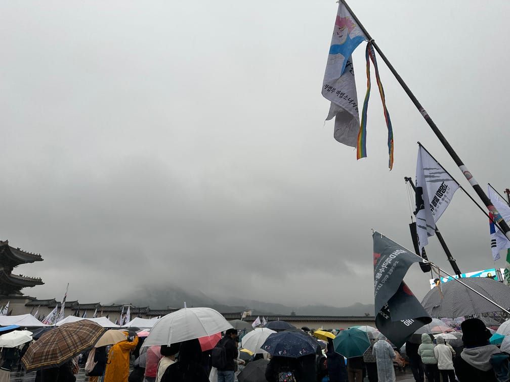 구름이 짙게 낀 경복궁 앞, 사람들이 우산을 쓰고 깃발을 들고 모여 있다. 한편에는 대형 전광판이 켜져 있는 것처럼 보인다.