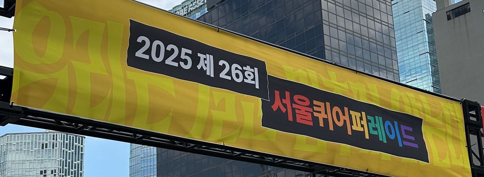 『2025 제 26회 서울퀴어퍼레이드』라고 적힌 현수막. 노란 배경과 글씨로 "우리는 결코 멈추지 않는다"라는 문구가 적혀 있다.