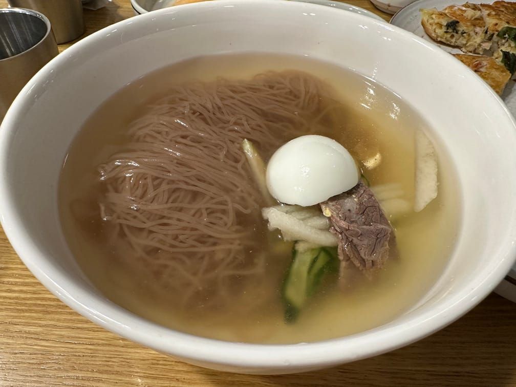 그릇에 메밀면, 맑은 국물, 수육, 채썬 오이와 무절임과 배, 달걀 반쪽이 차려져 있다.