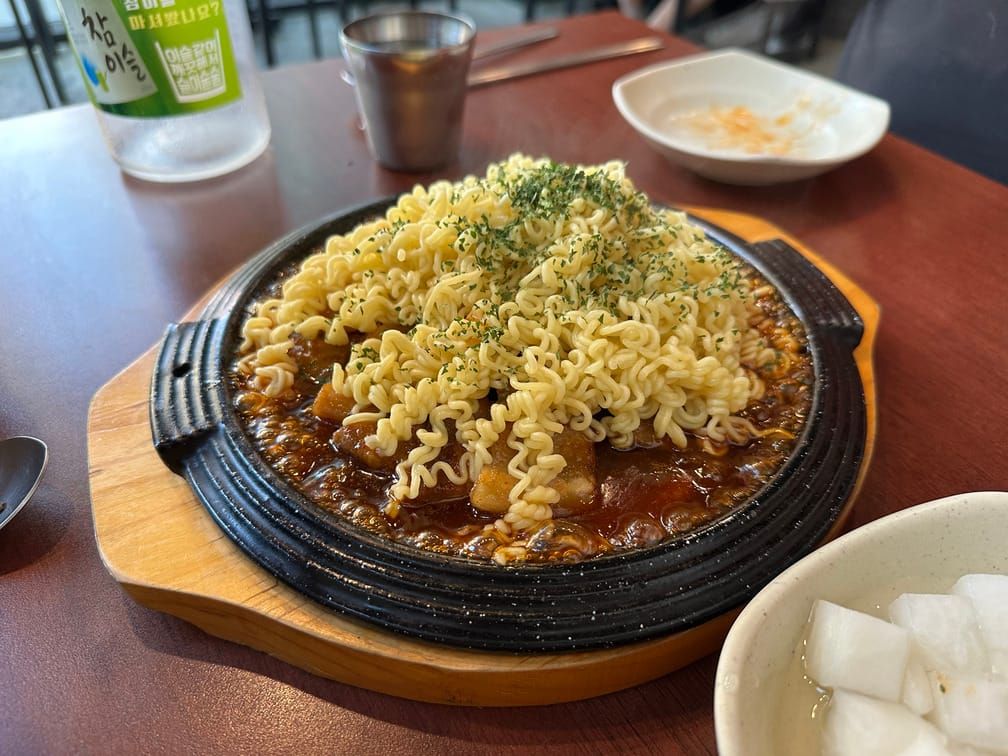 동그란 철판구이 그릇 안에 붉은 소스 삼겹살과 라면, 파슬리가 차려져 있다.
