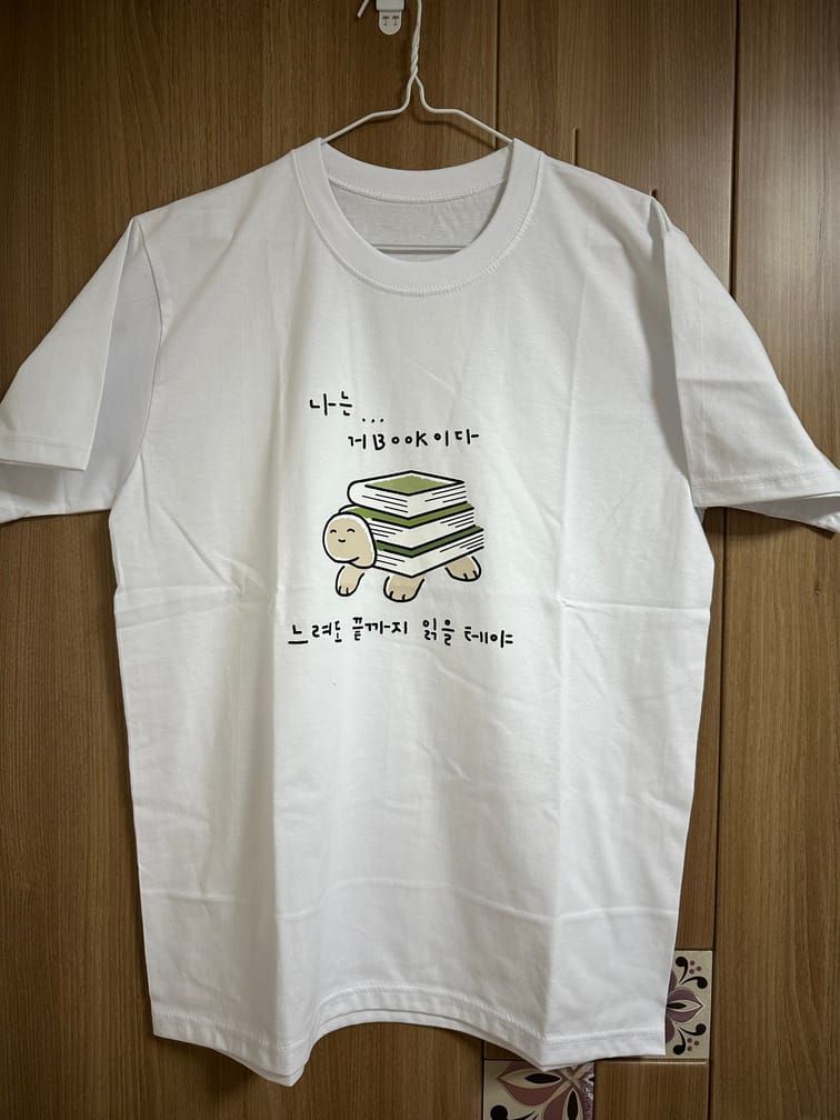 흰 티셔츠의 가슴 부분에 등껍질이 책 세 권으로 대체된 거북이 그림과 그 위아래로 "나는… 거BOOK이다", "느려도 끝까지 읽을 테야"라는 필기체 문장이 있다.